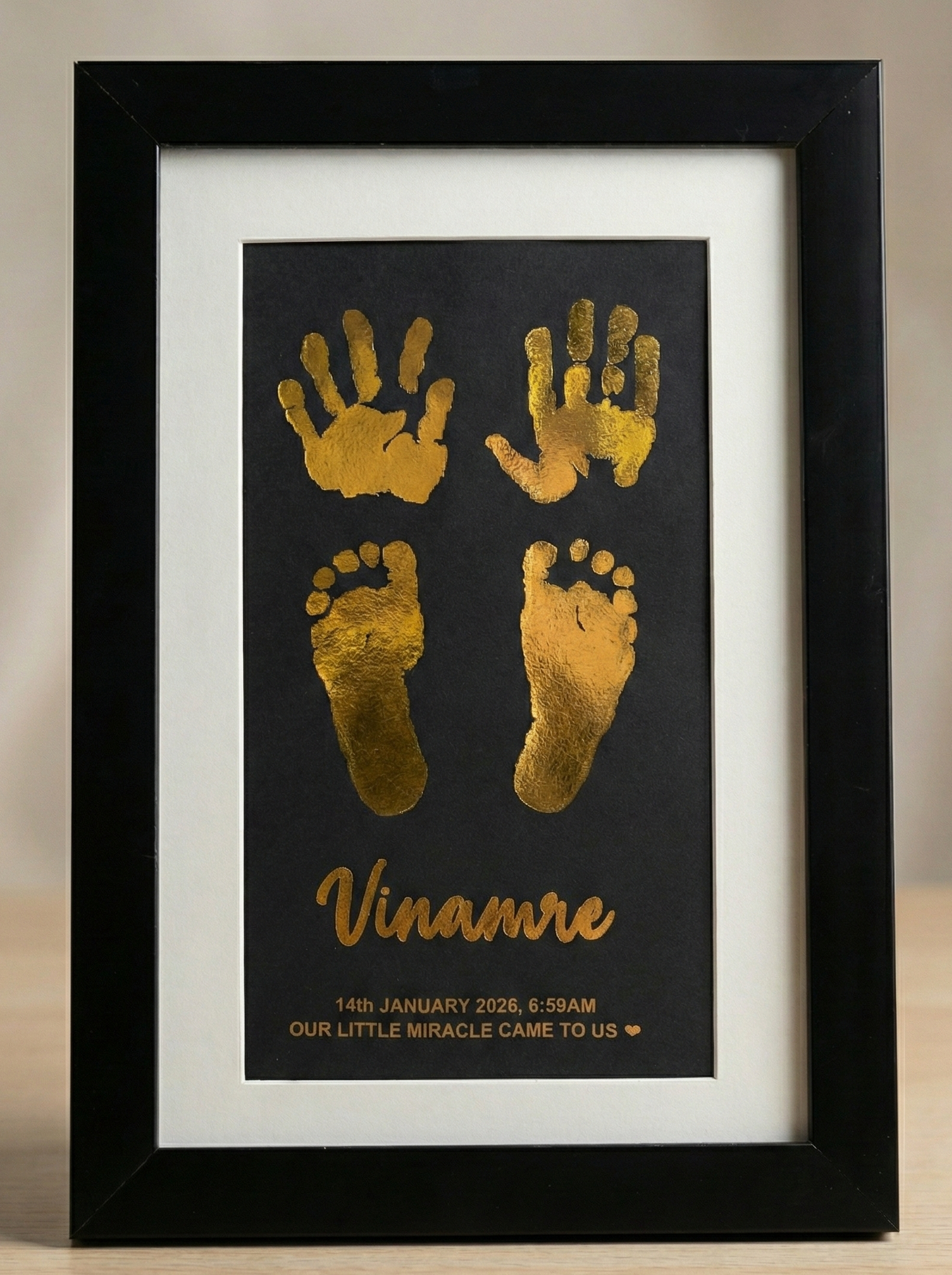 Framed artwork with gold hand and footprints and the name on a black background.
