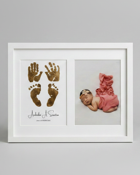 A white square framed keepsake print featuring gold baby footprints and handprints on the left, and a small photograph of a sleeping newborn baby in a pink outfit on the right, set against a neutral gray background.
