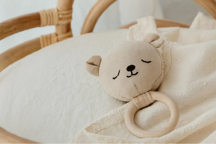 A soft baby sensory toy set in a natural rattan bassinet, featuring two star-shaped fabric cushions hanging from a wooden ring and a plush bear rattle resting on the cream bedding.