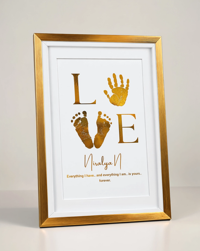 A framed personalized baby keepsake print featuring gold foil hand and footprints spelling 'LOVE' with the name 'Niralya N' and a quote below, in a gold frame against a neutral light gray background.