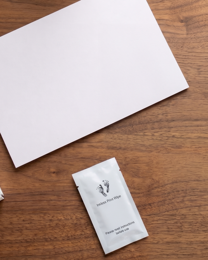 A small white sachet labeled 'Inkless print wipe' with a footprint logo, placed on a wooden tabletop next to a large blank white sheet.