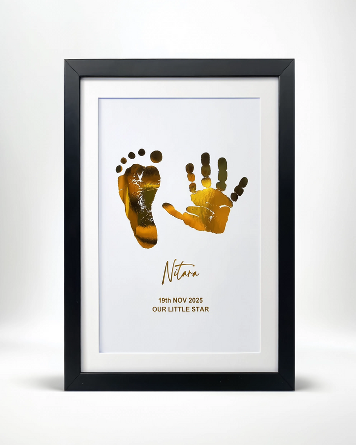 A framed personalized baby keepsake print featuring gold footprints and handprints on a white background, with the name 'Natura' and the date '19th NOV 2025 OUR LITTLE STAR' printed below. The print is housed in a white mat and a sleek black frame, set against a clean white background.