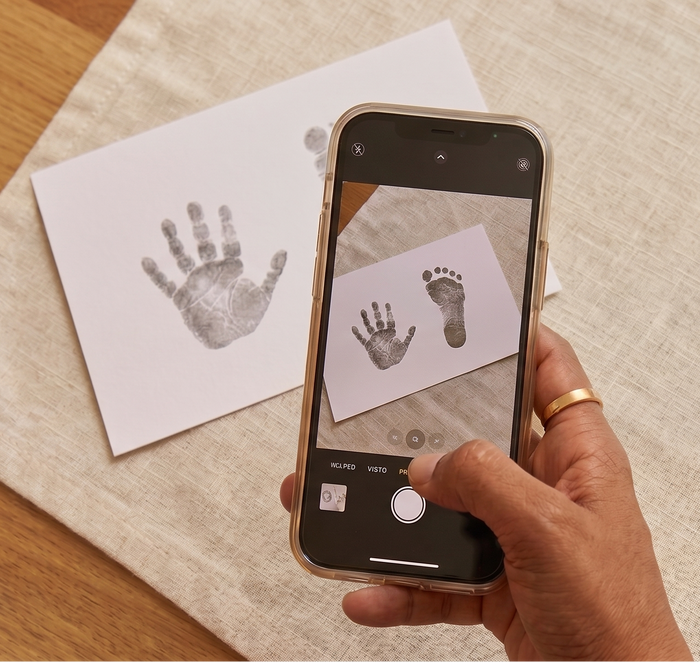 A hand holding a smartphone with a camera app open, showing a photo of baby footprints on a small card. A matching printed baby footprint card lies on a beige linen cloth on a wooden table.