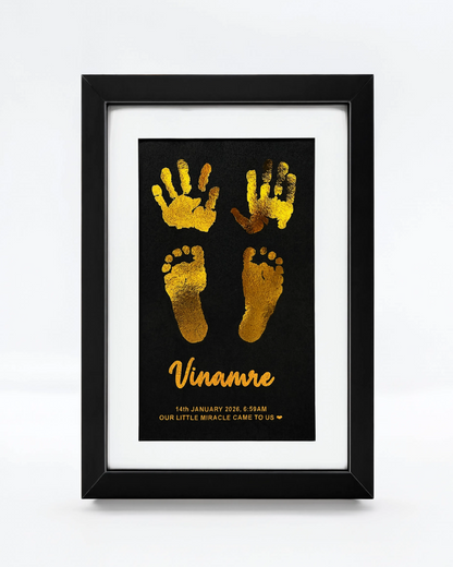 A framed personalized baby keepsake print featuring gold foil handprints and footprints on a black background, with the name 'Vinamre' in gold script. The print is matted in white and housed in a black frame, shown against a clean white background.