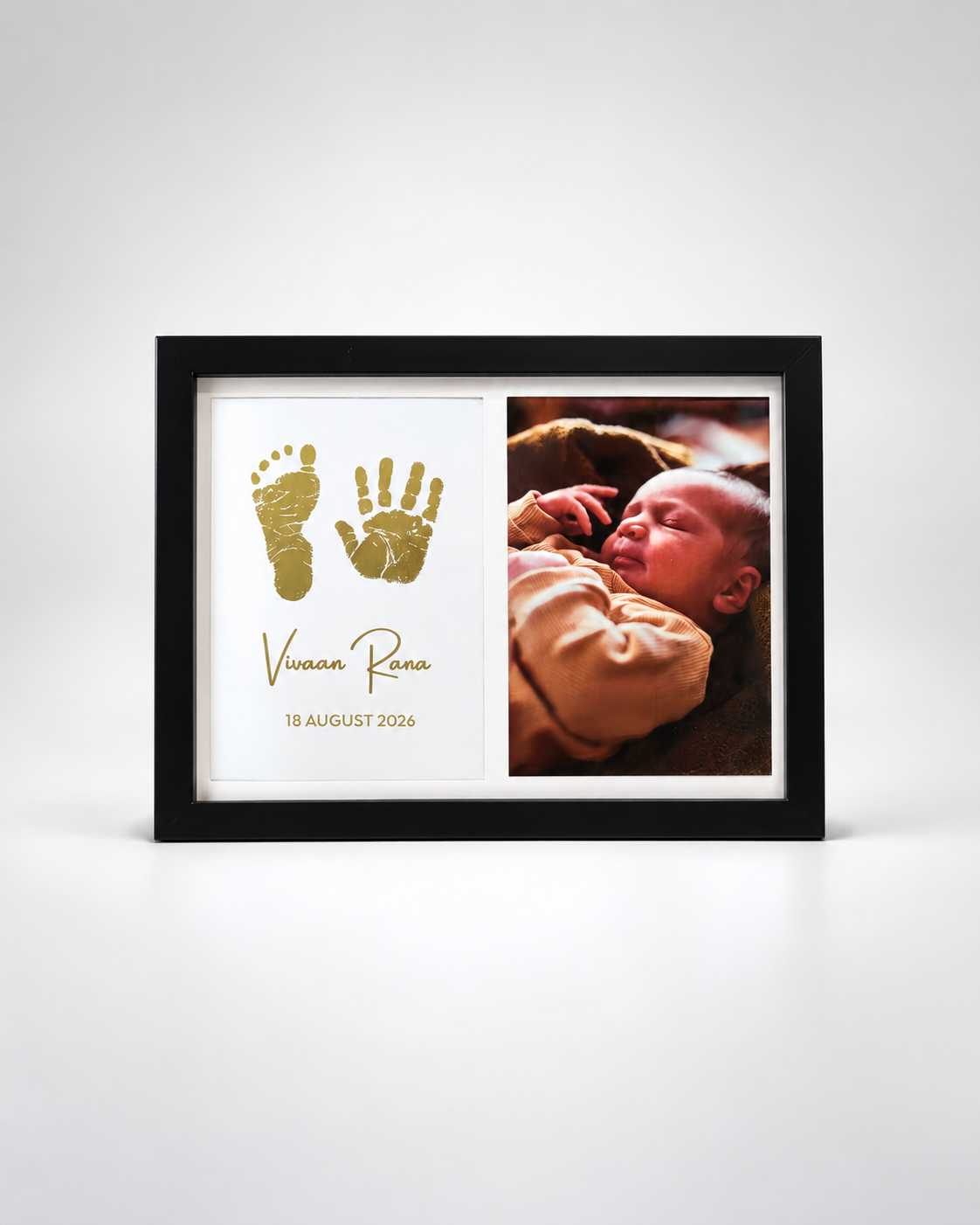A black-framed personalized baby keepsake photo frame on a light gray background. The left side features a white panel with gold foil baby footprints and handprints, the name 'Vivian Rona' in gold script, and the date '18 AUGUST 2026'.
