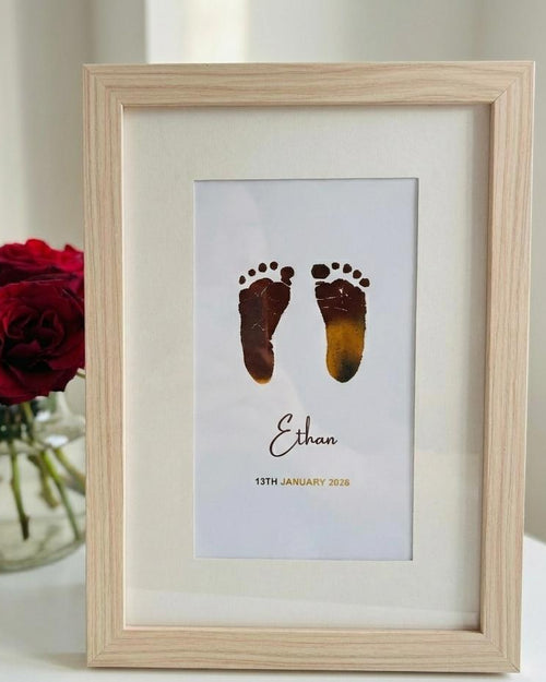 Framed print with baby footprints and name 'Ethan' next to red roses on a white surface