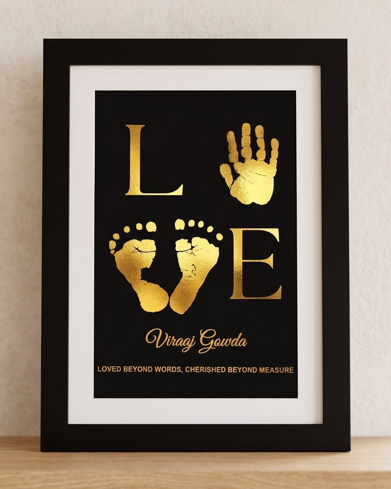 A framed personalized baby keepsake print featuring the word 'LOVE' with a handprint for the 'O' and baby footprints for the 'V' and 'E'. The print has a black background with gold foil lettering and is housed in a black frame with a white mat, leaning on a light wood shelf against a textured beige wall.