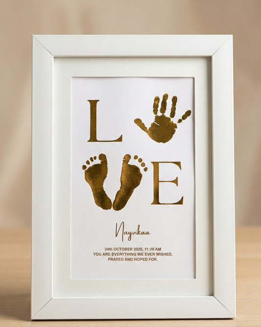 A white framed personalized keepsake print featuring gold foil handprints and footprints spelling LOVE, with a child's name and a short memorial message below, displayed on a light wood surface against a soft beige background.